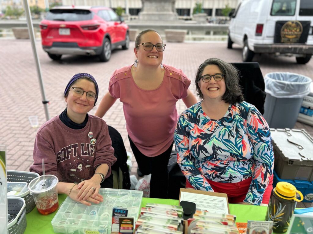 Three smiling lirarians sit under a tent behind a table ready to welcome visitors
